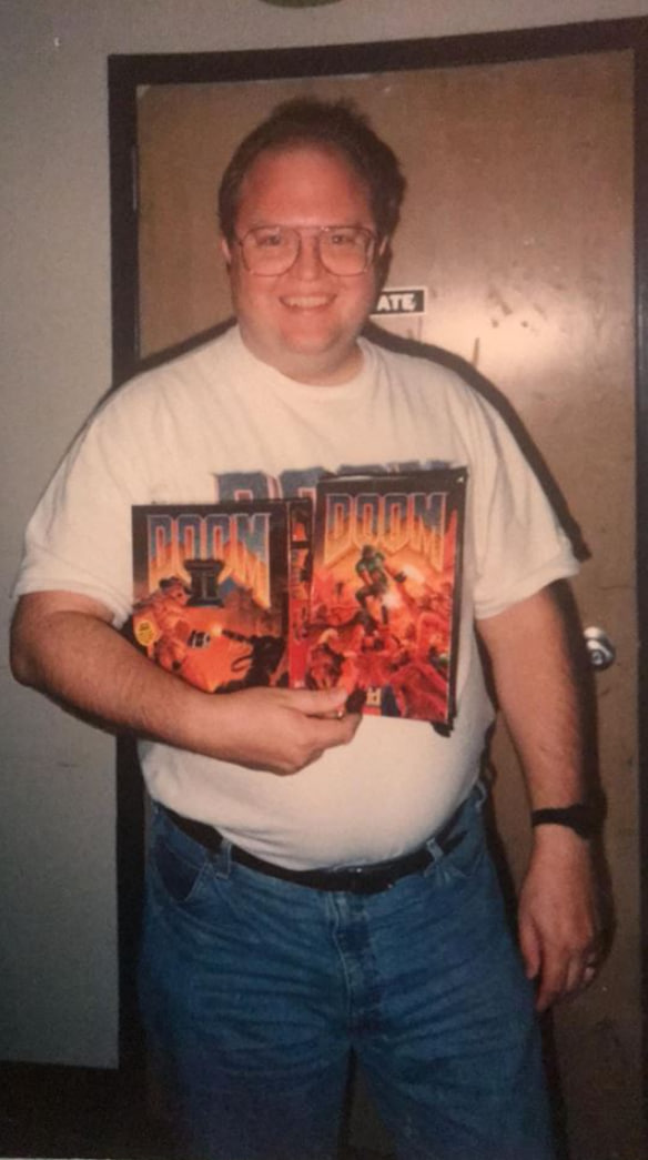 Sandy Petersen in the early 1990s, smiling and holding boxed copies of DOOM and DOOM II, wearing a DOOM II t-shirt.